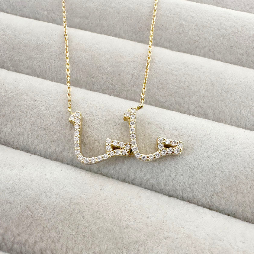 Close-up of a gold necklace with the word “ماما” (Mama) in Arabic script, encrusted with diamonds, resting on a soft beige jewelry display.