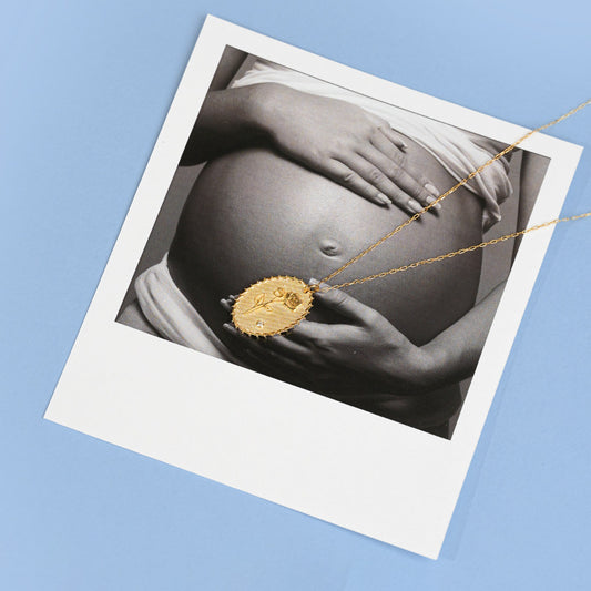 Gold necklace with a pendant over a black and white photo of a pregnant belly.