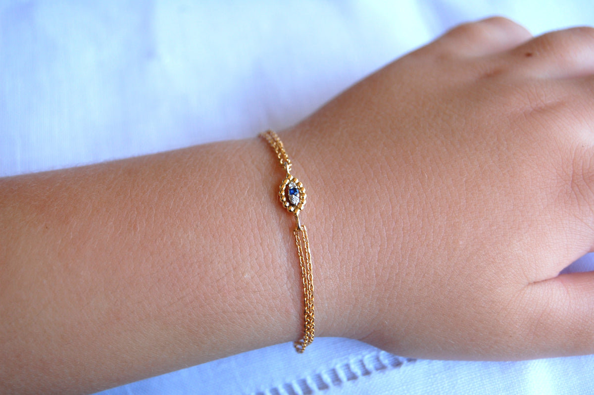 A close-up of a delicate 18K gold evil-eye bracelet worn on a wrist, featuring an oval of gold beads surrounding a central blue sapphire and two small diamonds, suspended on a fine double gold chain.