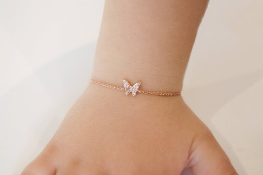 A close-up of a child’s wrist wearing a delicate rose-gold butterfly bracelet: the tiny butterfly pendant’s wings are pavé-set with sparkling diamonds, suspended on a fine adjustable rose-gold chain.