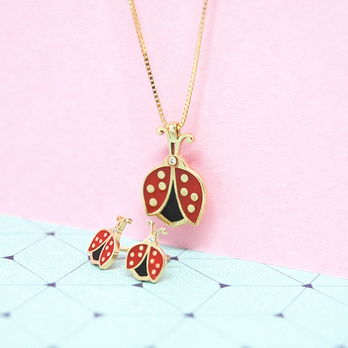 Matching ladybug necklace and earrings set in red, black, and gold, styled on a pastel pink background with a light blue patterned surface.
