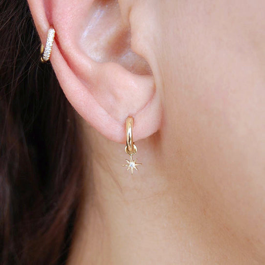 A close-up of a womanβs ear wearing the Mini Stellar Hoop: a small yellow-gold hoop from which dangles a delicate eight-point star with a sparkling diamond center.