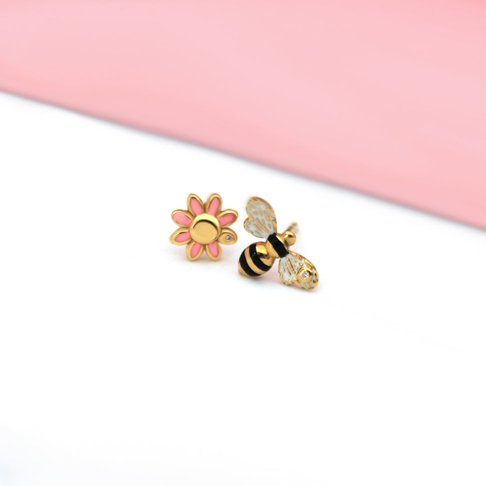 Mismatched stud earrings featuring a pink enamel daisy with a diamond center and a black, white, and gold enamel bee with a diamond on its wing, styled on a soft pink and white background.