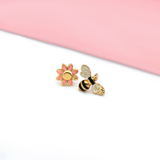 Mismatched stud earrings featuring a pink enamel daisy with a diamond center and a black, white, and gold enamel bee with a diamond on its wing, styled on a soft pink and white background.