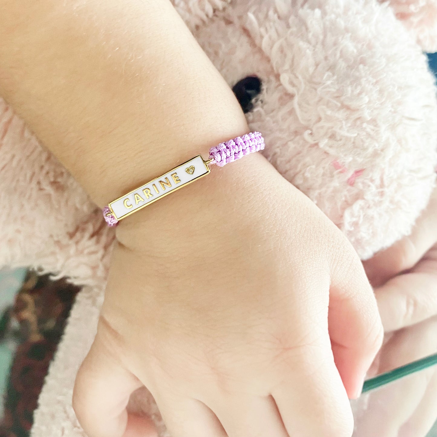 A close-up of a baby’s wrist wearing the BabyChic Enamel Name Bar Baby Bracelet in lavender: a pink braided cord with a white-enamel gold bar stamped ‘CARINE’ and a small gold heart, secured with an adjustable sliding knot.