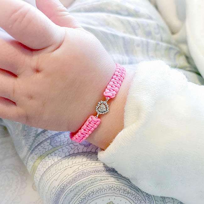 Pink woven bracelet with a heart-shaped white gold and diamond charm worn on a baby’s wrist, shown in a cozy indoor setting.