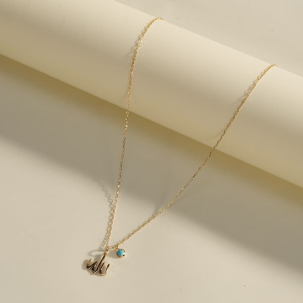 Gold necklace with “Allah” Arabic calligraphy pendant and a turquoise charm lying on a cream surface in soft daylight.