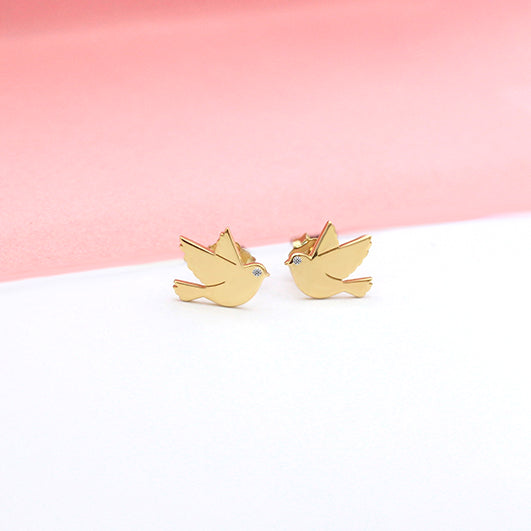 Pair of gold bird silhouette stud earrings with diamond eyes, placed on a soft white and pink background.