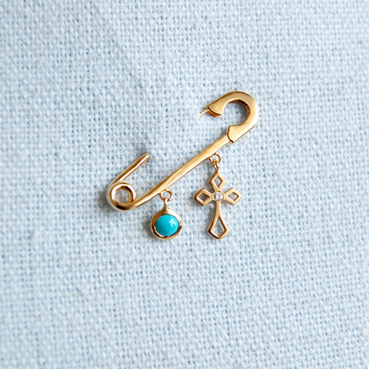 A polished 18K yellow-gold safety-pin brooch fastened into pale gray star-printed fabric, with two dangling charms: a round bezel-set turquoise cabochon and an open Arabic ‘Allah’ script pendant.