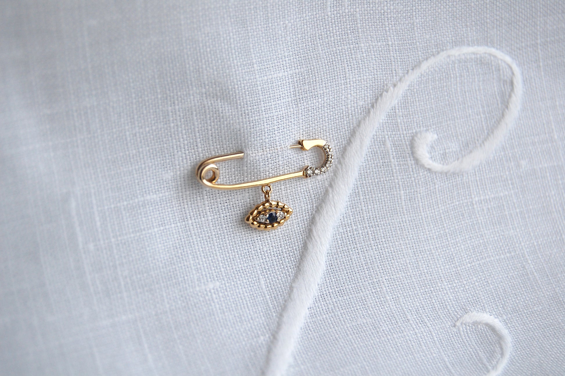A close-up styling shot of an 18K yellow-gold safety-pin brooch resting on white linen, with the pin’s clasp tip set with small diamonds and a dangling evil-eye charm of gold beads around a central blue sapphire and two diamonds.