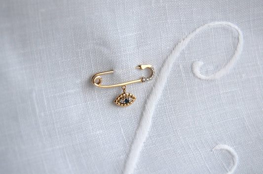 A close-up styling shot of an 18K yellow-gold safety-pin brooch resting on white linen, with the pin’s clasp tip set with small diamonds and a dangling evil-eye charm of gold beads around a central blue sapphire and two diamonds.