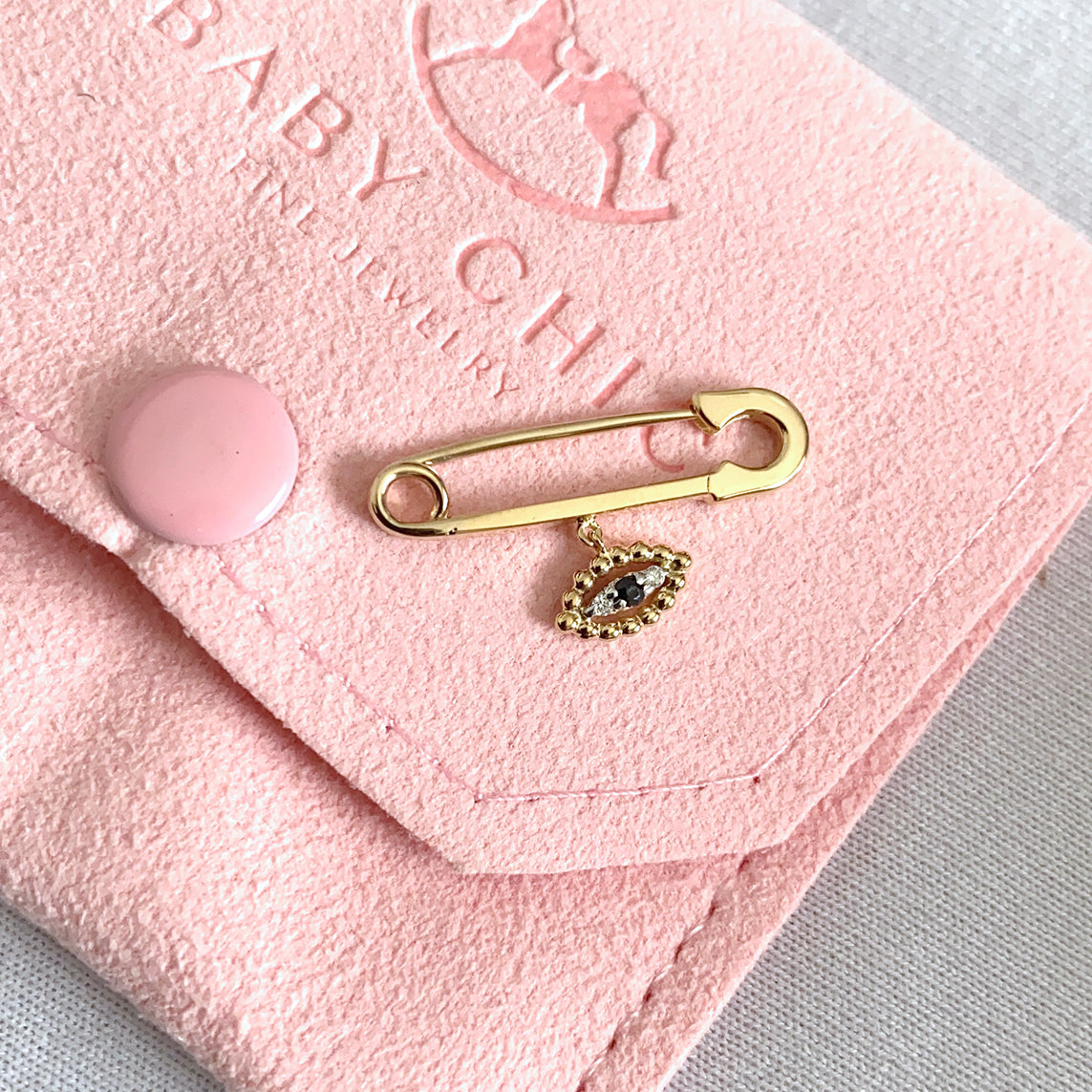 A polished 18K yellow-gold baby safety pin brooch with a dangling evil-eye charm—an eye-shaped cluster of gold beads around a central dark blue gemstone and two small diamonds—resting on a blush-pink suede pouch embossed with the BabyChic logo