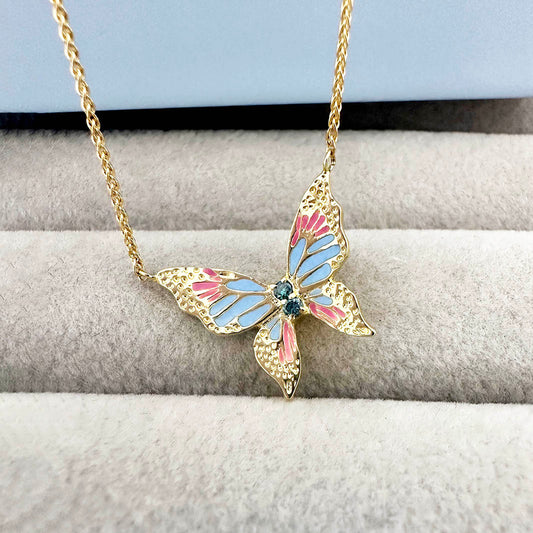 Close-up of a gold butterfly necklace with pink and blue enamel wings and three blue diamonds in the center, displayed on a soft jewelry tray.