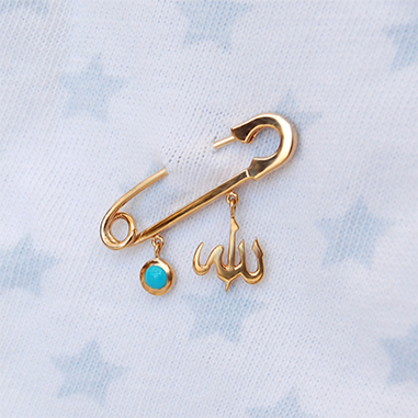 A polished 18K yellow-gold safety-pin brooch fastened into a white fabric printed with pale blue stars, suspending two little charms: a round turquoise enamel disc and an Arabic ‘Allah’ script pendant.