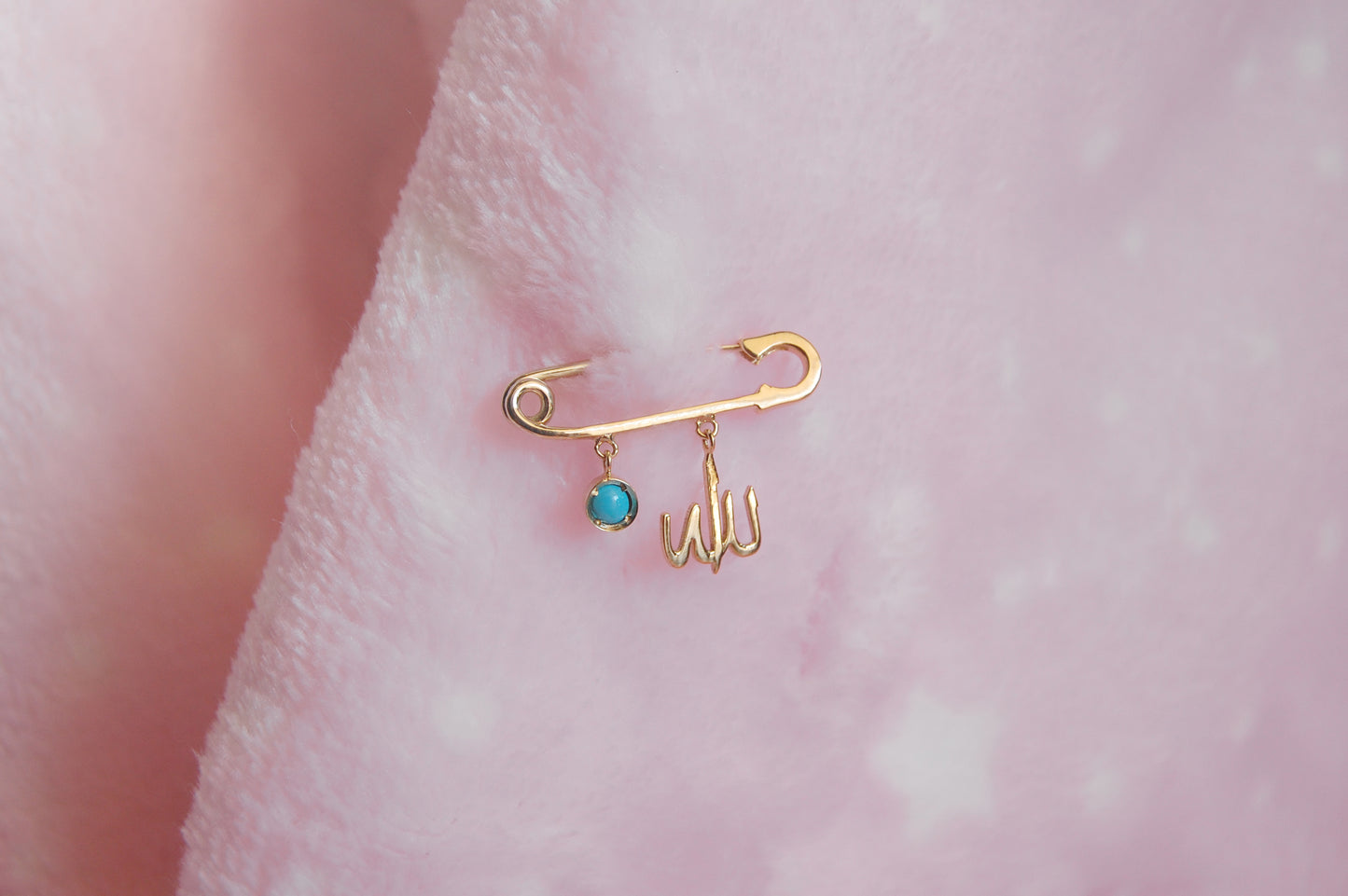 A polished 18K yellow-gold safety-pin brooch pinned into a soft pink plush fabric, with two dangling charms: a round turquoise-enamel disc and a delicate Arabic ‘Allah’ script.