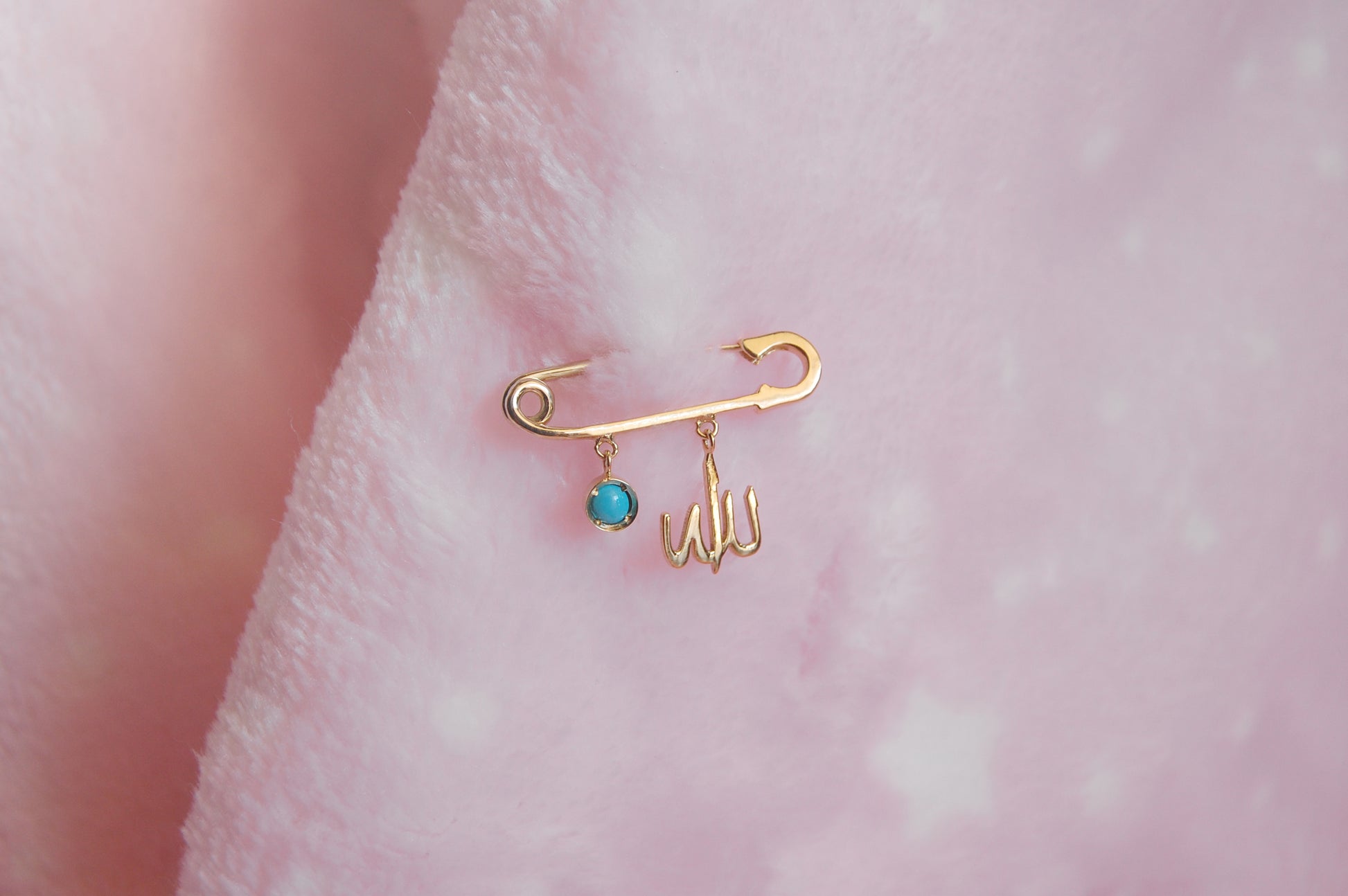 A polished 18K yellow-gold safety-pin brooch pinned into a soft pink plush fabric, with two dangling charms: a round turquoise-enamel disc and a delicate Arabic ‘Allah’ script.