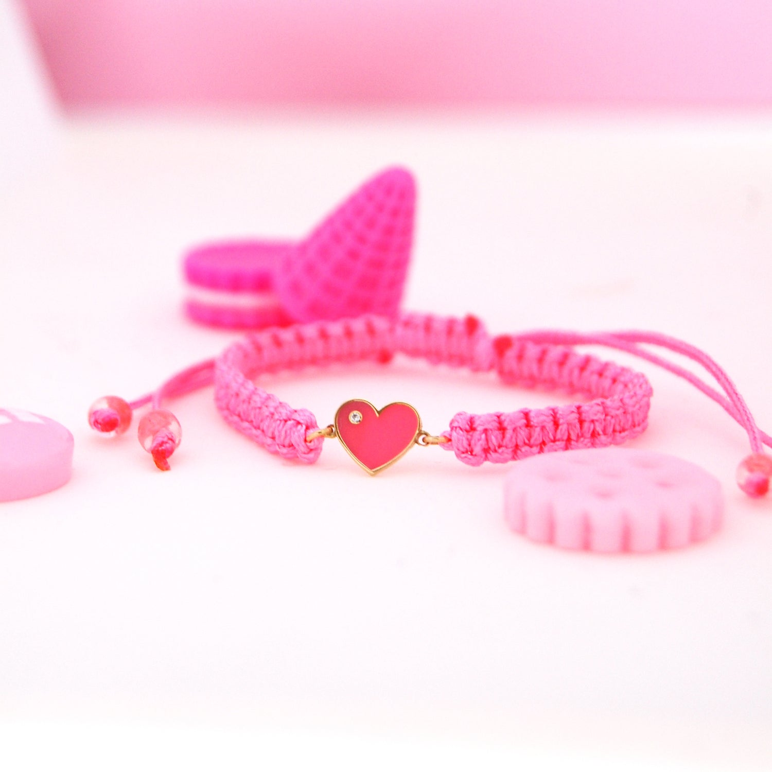 Flat-lay of the Forever Love Diamond Bracelet surrounded by pastel candy-like props, featuring a pink enamel heart with a diamond on a pink woven band.