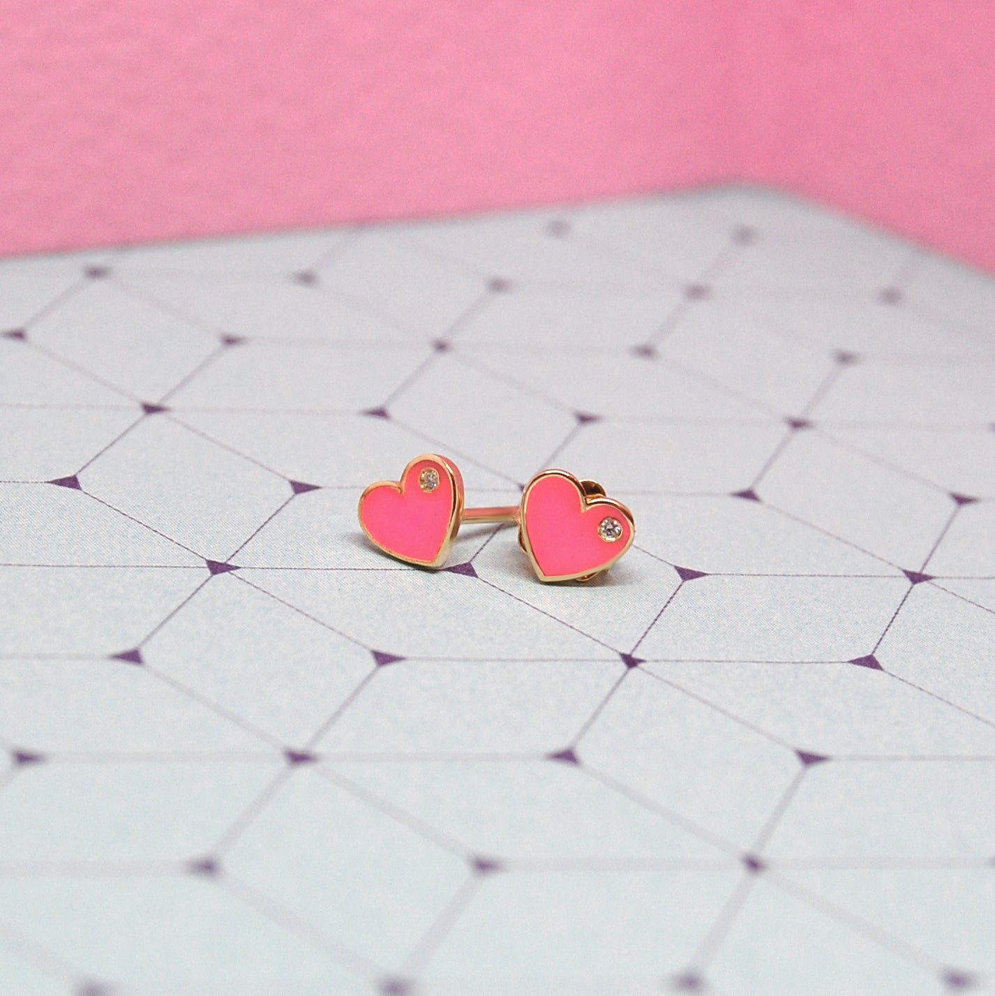 Pink heart-shaped enamel earrings with diamond accents displayed on a geometric purple-patterned background.