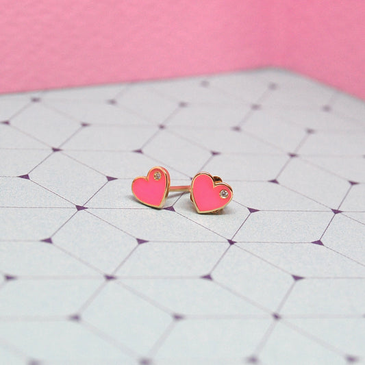 Pink heart-shaped enamel earrings with diamond accents displayed on a geometric purple-patterned background.