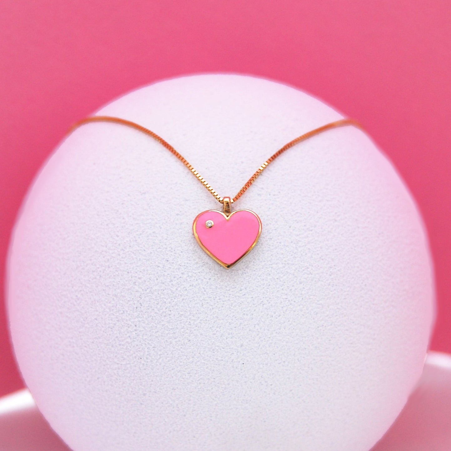 Pink heart-shaped pendant necklace with a diamond accent, displayed elegantly on a white textured stand against a pink background.