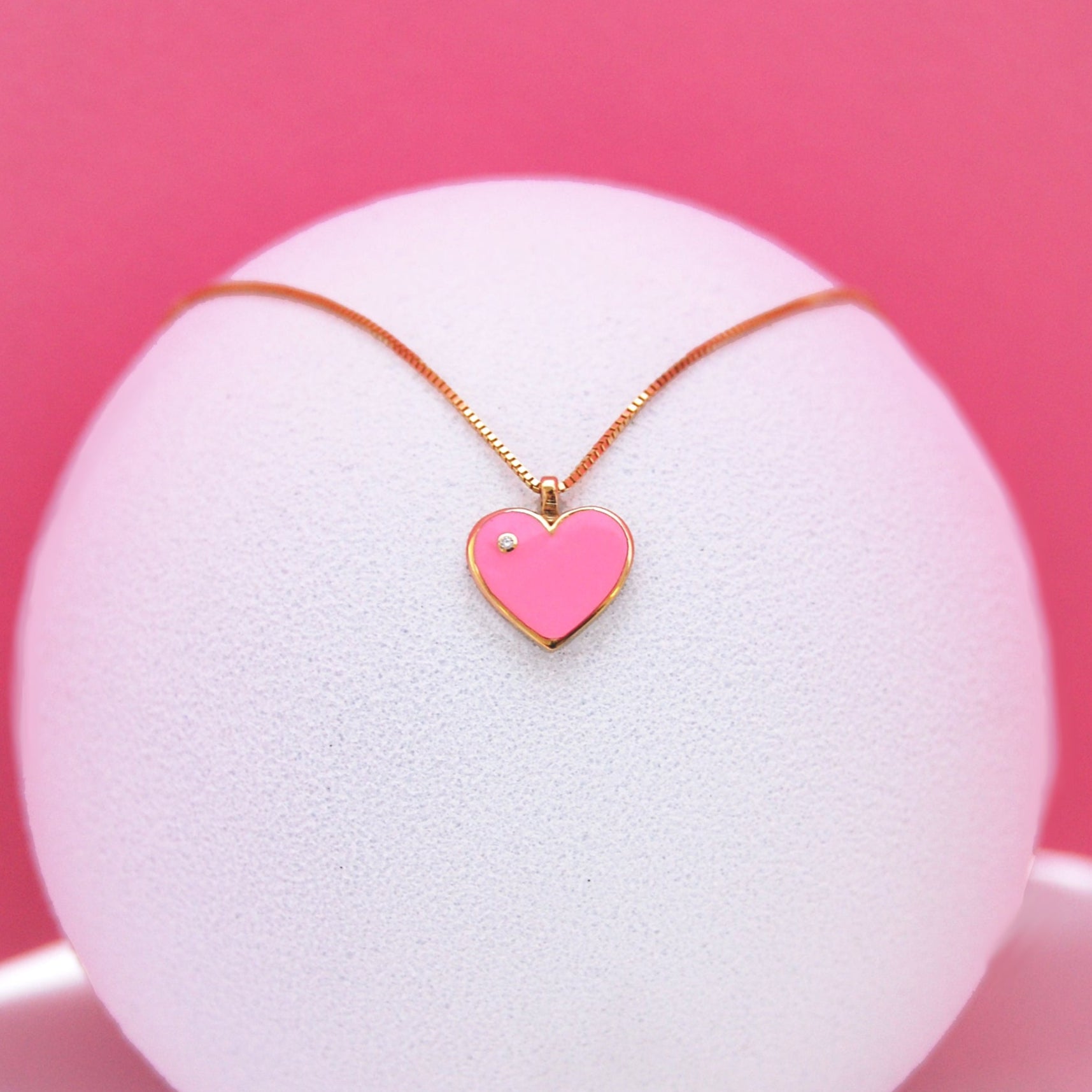 Pink heart-shaped pendant necklace with a diamond accent, displayed elegantly on a white textured stand against a pink background.