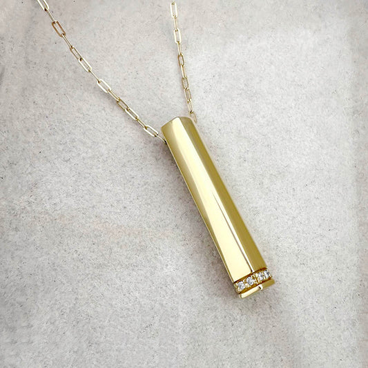 Gold vertical bar pendant necklace with diamond accents at the base, shown on a soft light grey jewelry display.