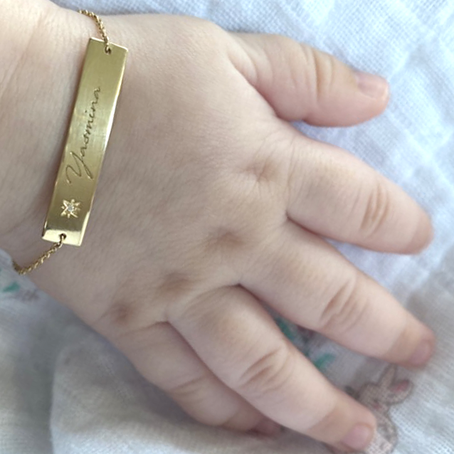 A close-up of a baby’s hand wearing the Inscription Diamond Bracelet: a slender 18K yellow-gold bar engraved with ‘Yasmina’ and accented by a small starburst diamond, suspended on a delicate chain around a tiny wrist.