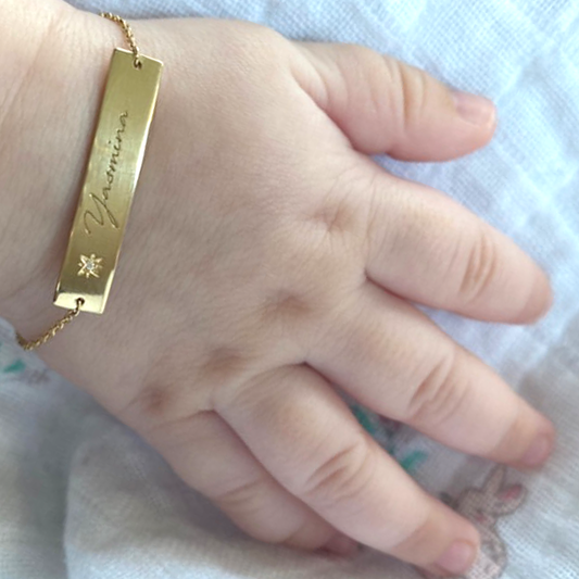 A close-up of a baby’s hand wearing the Inscription Diamond Bracelet: a slender 18K yellow-gold bar engraved with ‘Yasmina’ and accented by a small starburst diamond, suspended on a delicate chain around a tiny wrist.