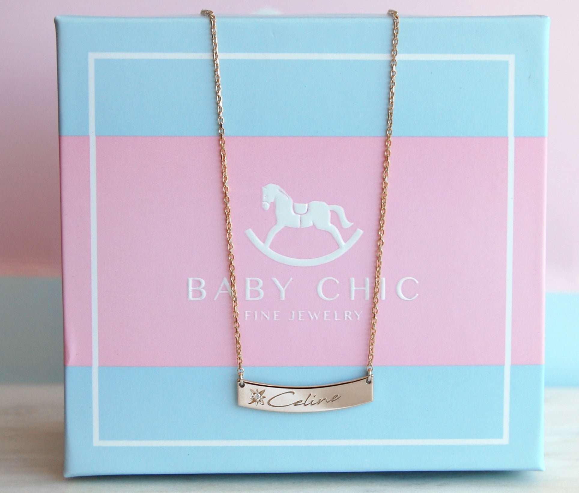 A delicate 18K yellow-gold curved bar pendant engraved with the name “Celine” and featuring a small starburst motif set with a round diamond, suspended from a fine paper-clip chain and displayed on a pastel blue and pink BabyChic jewelry box.