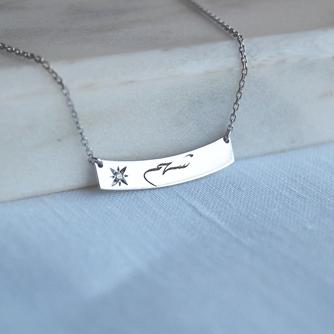 A close-up of a personalized white-gold curved bar necklace resting on pale gray linen, the bar engraved in flowing Arabic script beside a starburst motif set with a small round diamond, suspended from a fine paper-clip chain.