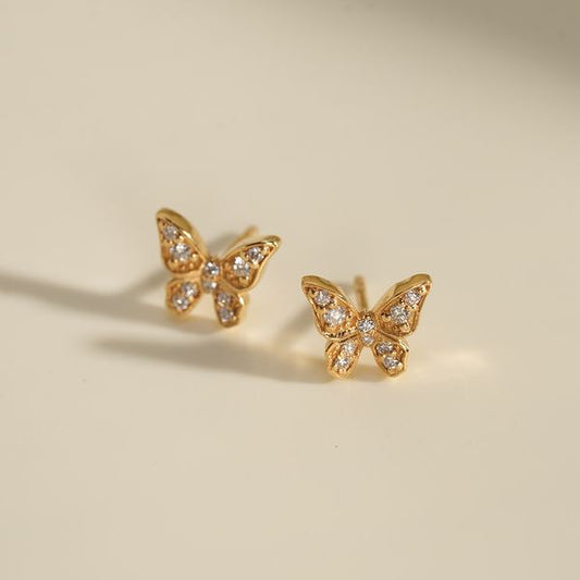 Little Butterfly Earrings – delicate 18K gold studs shaped like butterflies, encrusted with tiny diamonds, softly glowing against a cream background.