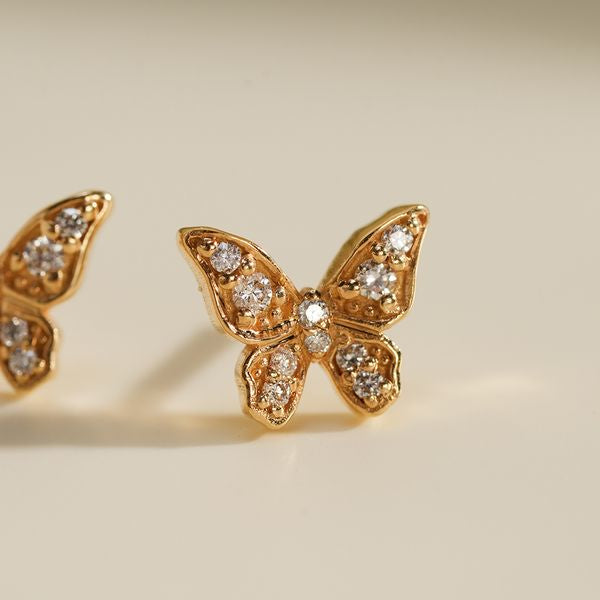 Close-up of a single Little Butterfly Earring in 18K gold, adorned with sparkling diamonds on each wing, glowing softly against a cream background.