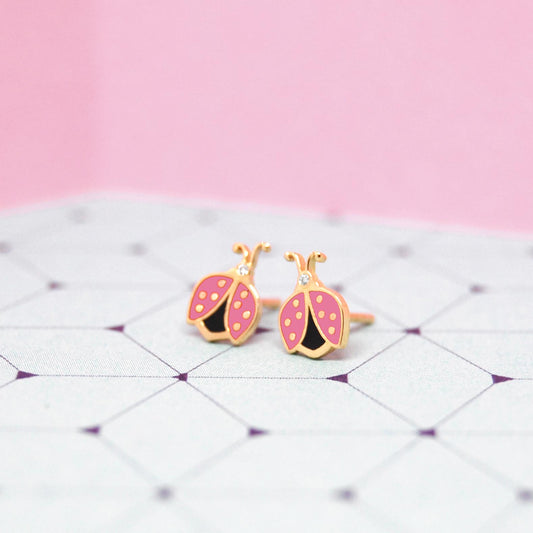 Pink enamel ladybug earrings with gold and black detailing, displayed on a geometric lavender surface with a soft pink background.