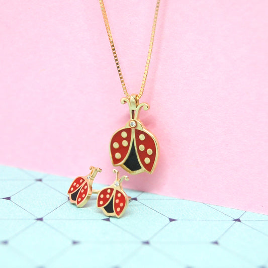 Close-up of gold ladybug pendant necklace with matching earrings, shown against a pastel pink and blue geometric background.