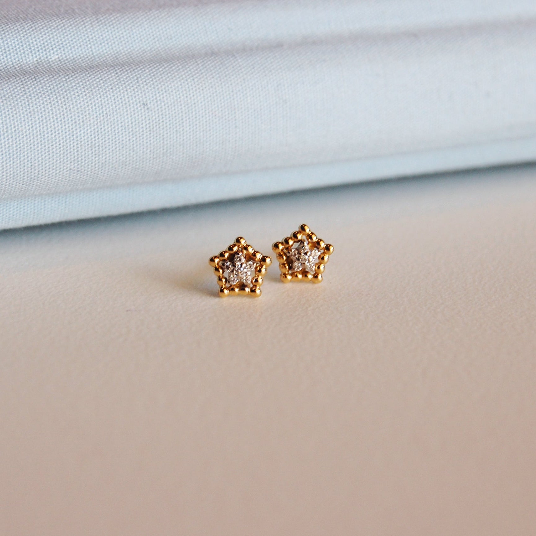 Pair of Little Star Earrings in gold, featuring a pavé diamond center, set on a soft cream surface with a pastel background.