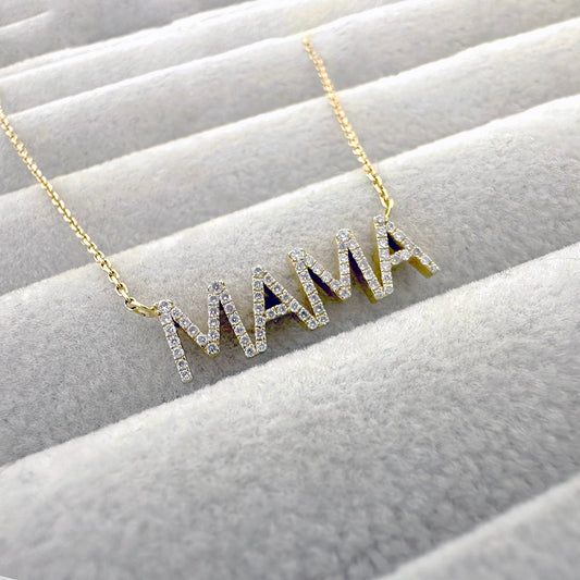 Gold “MAMA” necklace with diamond-studded capital letters, displayed on a soft beige jewelry tray.