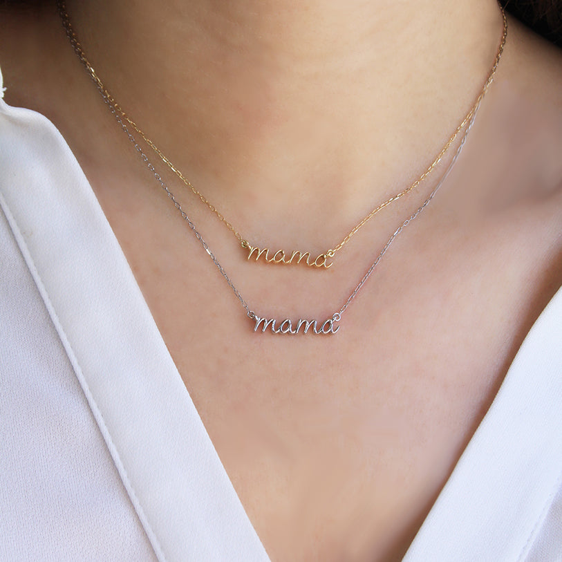 Two delicate “mama” script necklaces worn layered on the neck—one in yellow gold and one in white gold—displayed flat against skin and a white blouse.