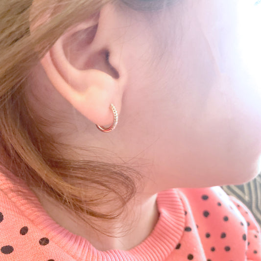 A close-up of a woman’s ear wearing a delicate 18K yellow-gold mini hoop earring studded with a row of small round diamonds along the front half of the hoop.