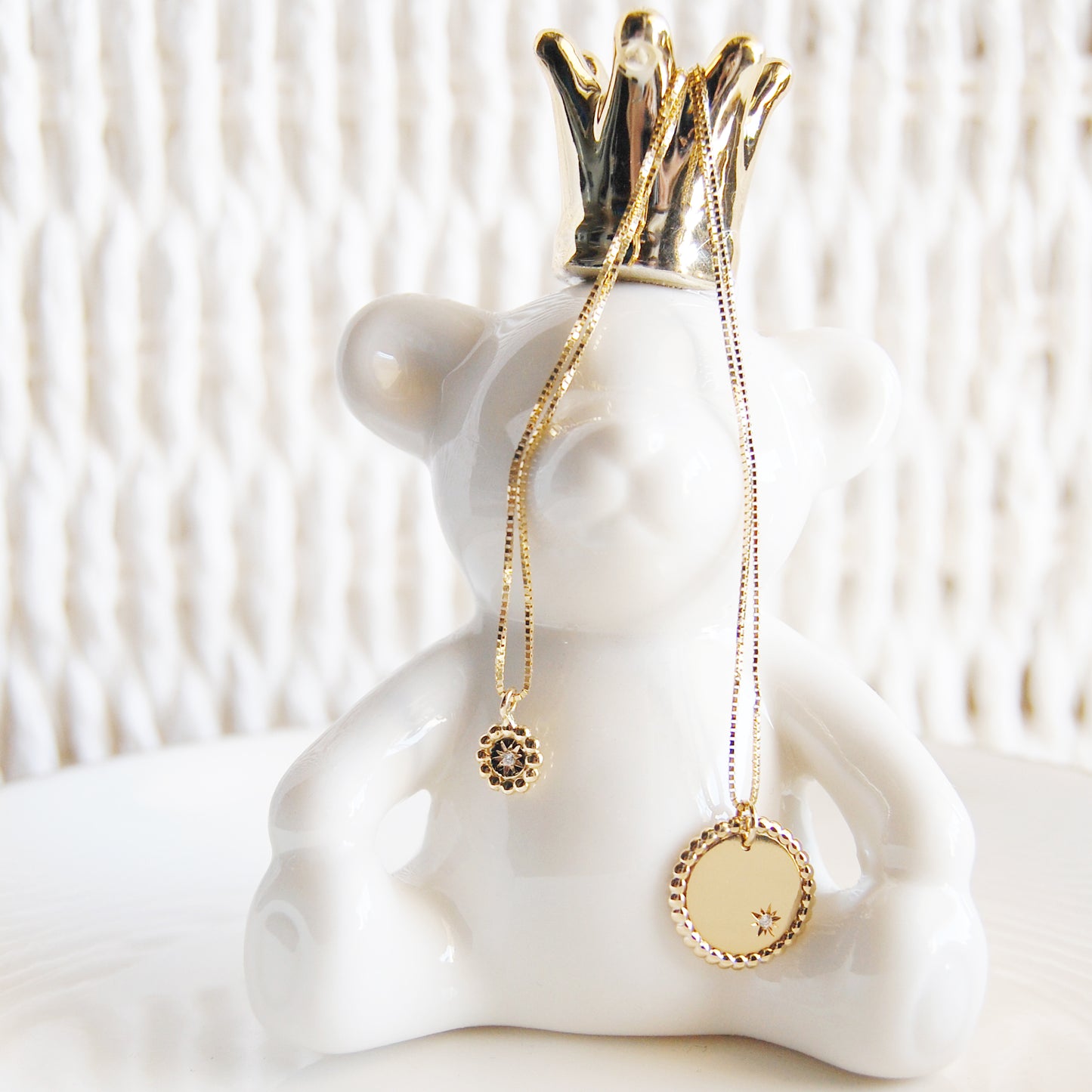 A white ceramic bear figurine wearing a gold crown, draped with two yellow-gold necklaces: on the left, a mini scalloped-bead coin pendant set with a single diamond; on the right, a larger scalloped-edge coin pendant engraved with a starburst and diamond—both hanging from fine paper-clip chains.