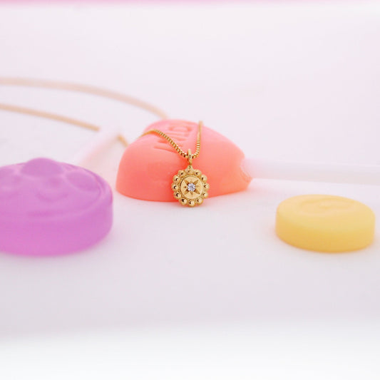 Mini Stellar Coin Necklace styled on pastel candy background, showcasing a round 18K gold pendant with beaded edges and a central diamond-set starburst.