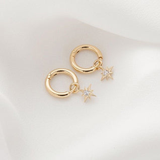A pair of 18K yellow-gold mini hoop earrings laid flat on a neutral surface, each hoop suspending a small eight-point star charm set with a single sparkling diamond.