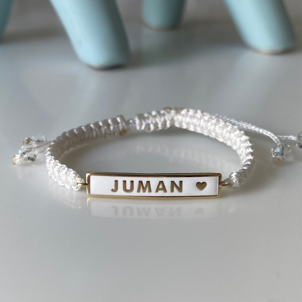 A BabyChic Enamel Name Bar Baby Bracelet with adjustable white braided cord, featuring a white-enamel gold-edged nameplate stamped ‘JUMAN’ alongside a small gold heart, resting on a glossy surface.