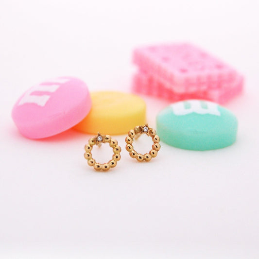 A pair of whimsical gold beaded earrings shaped in a circle with a tiny star and diamond accent at the top, styled against colorful candy pieces.