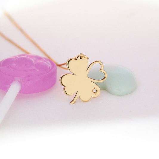 A close-up of the Piece of My Heart necklace pendant resting against pastel decor: an 18K yellow-gold clover-shaped charm formed by three solid heart-shaped leaves and one open-heart leaf, accented with a starburst motif and single diamond, shown on a fine chain with soft purple and mint props in the background.
