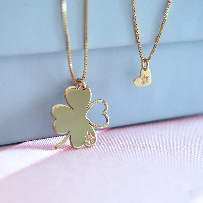 A close-up of two layered 18K yellow-gold necklaces displayed against pastel props: the longer chain holds a clover-shaped ‘Piece of My Heart’ pendant with three solid heart leaves and one open-heart leaf, accented by a small diamond starburst; the shorter chain carries a tiny heart pendant also set with a single diamond.