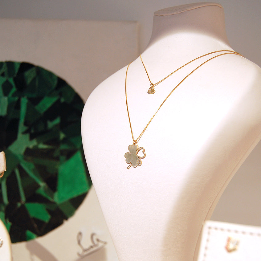 A display bust wearing two delicate 18K yellow-gold necklaces: the shorter chain holds a small heart-shaped pendant, and the longer chain features the ‘Piece of My Heart’ clover pendant with three solid and one open heart-shaped leaves.