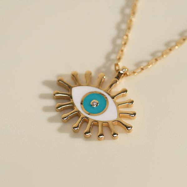 A close-up styling shot of the BabyChic Radiant Eye Diamond Necklace: the gold enamel eye pendant with white and turquoise enamel iris and a central diamond rests on a soft cream paper backdrop, its fine gold box chain forming a subtle shadow.