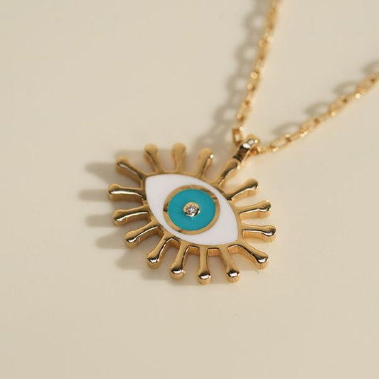 A close-up styling shot of the BabyChic Radiant Eye Diamond Necklace: the gold enamel eye pendant with white and turquoise enamel iris and a central diamond rests on a soft cream paper backdrop, its fine gold box chain forming a subtle shadow.