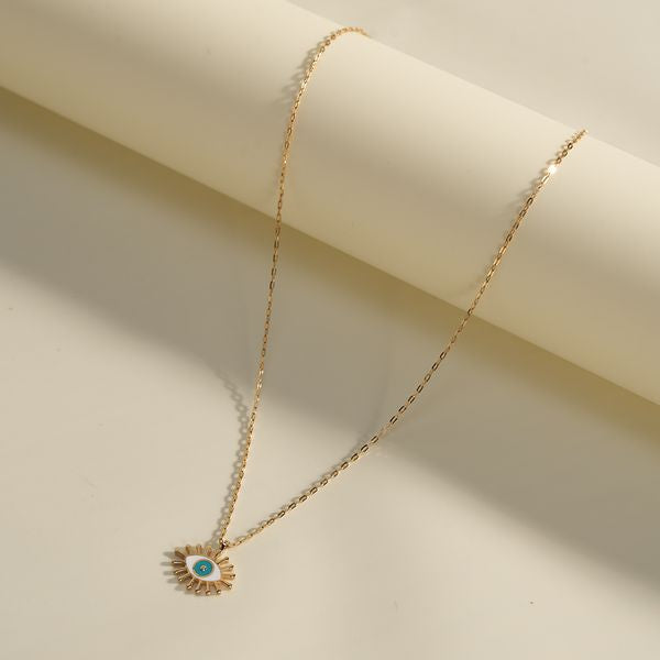 A BabyChic Radiant Eye Diamond Necklace styled against a cream-colored rolled paper backdrop: a fine 18K gold box chain drapes from the top edge, suspending an enamel eye pendant with white and blue iris, a central sparkling diamond, and radiating gold rays.