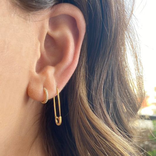 A close-up of a woman’s ear wearing two earrings: a petite 18K gold huggie hoop studded with tiny diamonds in the upper lobe, and the single-pin diamond earring—a safety-pin–shaped gold stud with diamonds along its curved clasp tip—in the lower lobe.